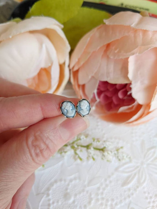 Blue Cameo earrings being held in fingers to show tiny size with pink flowers in the background