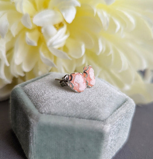 Pair of white on peach cameo earrings in silver settings on a hexagonal green velvet stand with white flowers in the background.