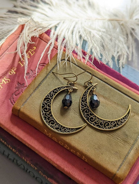 Antiqued Brass Filigree Crescent Moon Earrings with Black Teardrops on a textured surface with books and white feathers.