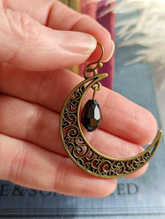 Antiqued Brass Filigree Crescent Moon Earring with Black Teardrop held between fingers with a blurred background