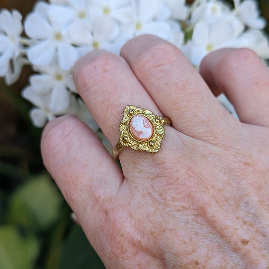 Gold Greek Goddess Cameo Ring - Shield Signet Setting holding a white on peach cameo on a hand in front of white flowers