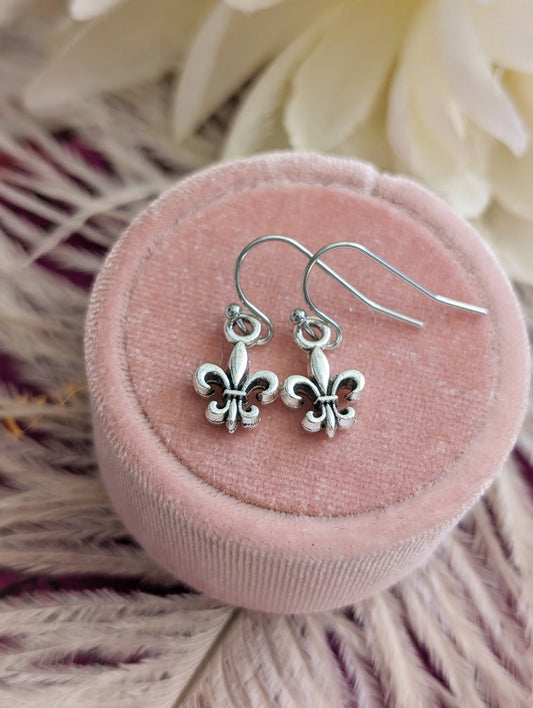 Silver fleur-de-lis earrings on a pink velvet cushion with white flowers in the background