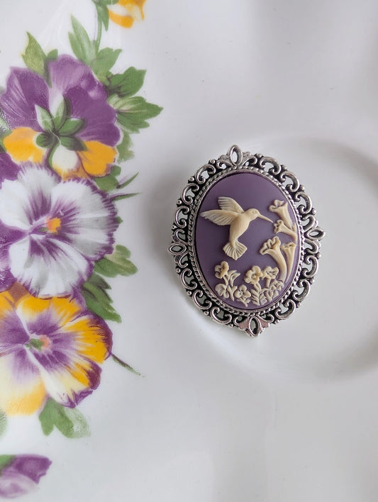 Silver lacy brooch featuring a cameo of a white hummingbird and trumpet flowers on a purple background. Displayed on a vintage plate with a floral motif.