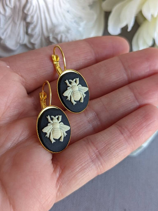 Pair of bee cameo earrings set in golden brass, sawtooth-edged lever back settings held in a hand against a neutral background