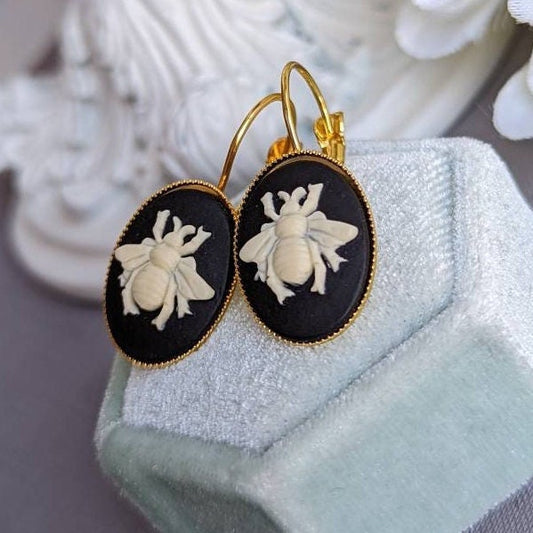 Pair of bee cameo earrings set in golden brass, sawtooth-edged lever back settings displayed on a pale green velvet jewelry box.