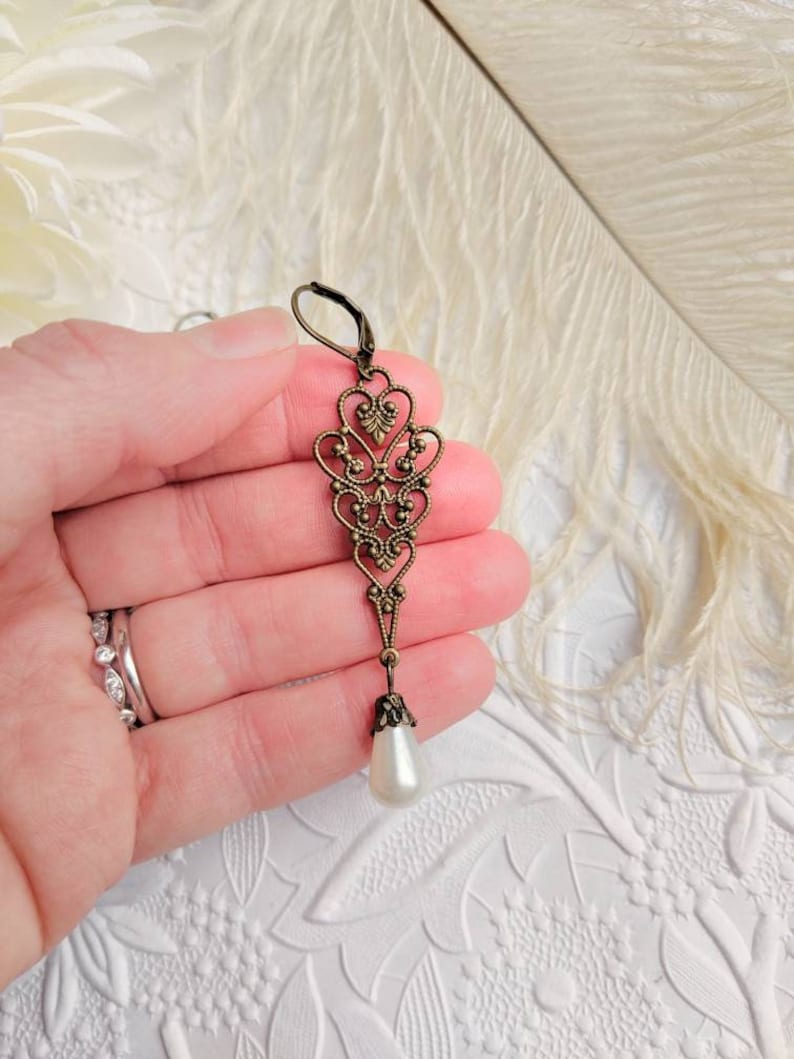Long Victorian Pearl Drop Earring - Bronze Filigree Bridal Dangle held in a hand against a textured white background