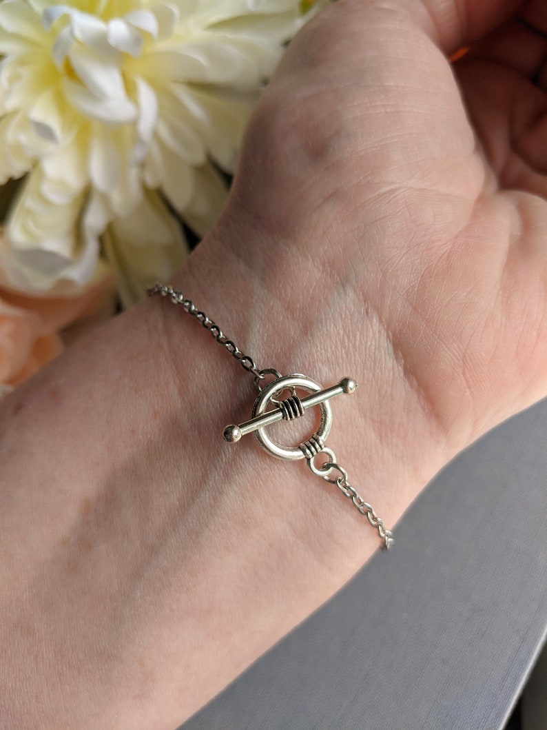 Silver bracelet with a toggle clasp on a wrist, with a blurred background of flowers.