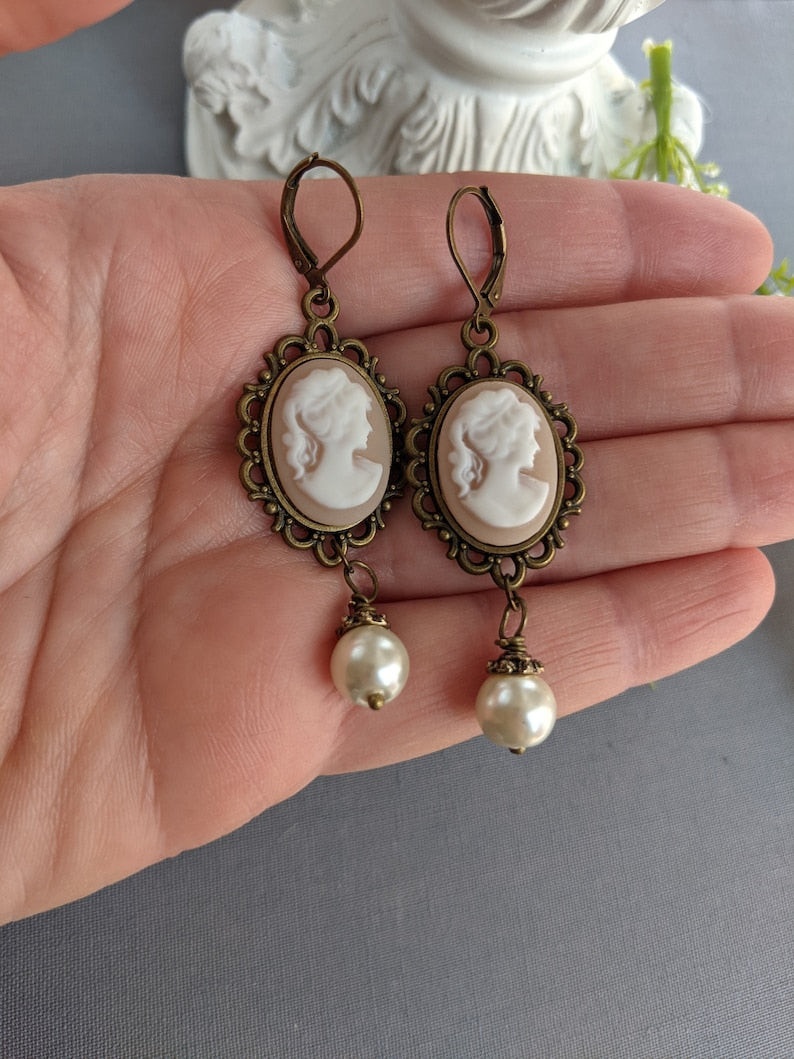 Pair of peach and white vintage lady cameo drop earrings with antiqued brass lace frames and glass pearl drops held in a hand against a neutral background.