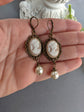 Pair of peach and white vintage lady cameo drop earrings with antiqued brass lace frames and glass pearl drops held in a hand against a neutral background.