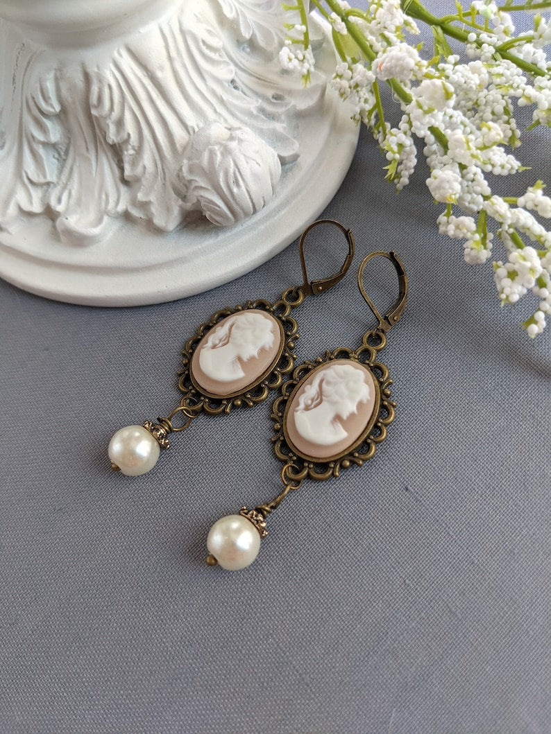 Pair of peach and white vintage lady cameo drop earrings with antiqued brass lace frames and glass pearl drops on a gray surface.
