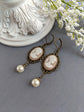 Pair of peach and white vintage lady cameo drop earrings with antiqued brass lace frames and glass pearl drops on a gray surface.