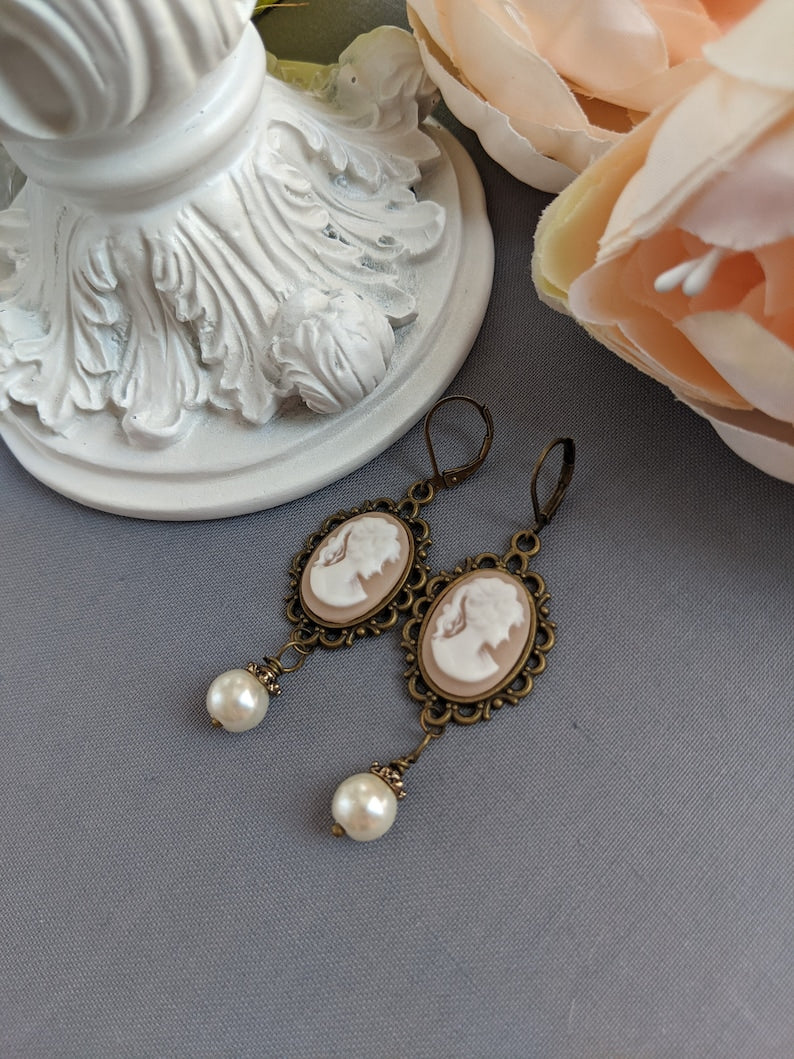 Pair of peach and white vintage lady cameo drop earrings with antiqued brass lace frames and glass pearl drops on a gray surface.