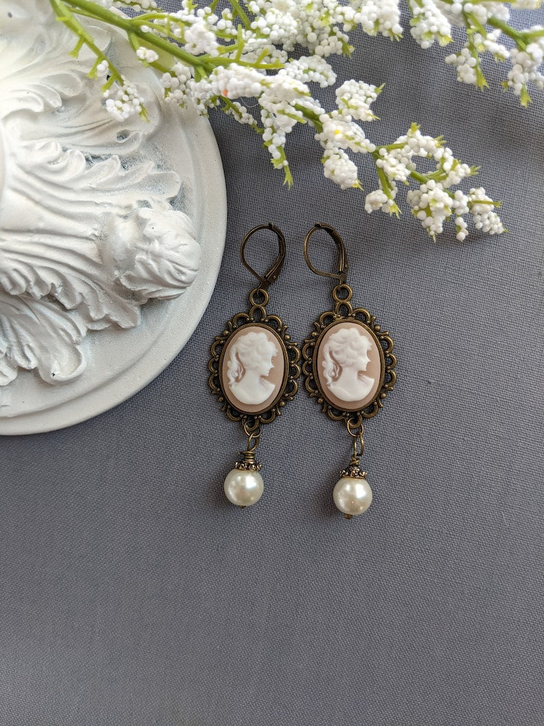 Pair of peach and white vintage lady cameo drop earrings with antiqued brass lace frames and glass pearl drops on a gray surface with white flowers.