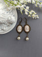 Pair of peach and white vintage lady cameo drop earrings with antiqued brass lace frames and glass pearl drops on a gray surface with white flowers.