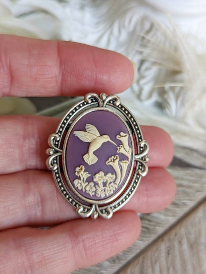 Silver flourish brooch featuring a cameo of a white hummingbird and trumpet flowers on a purple background. Shown being held in a hand.
