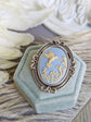 Silver flourish brooch featuring a cameo of a white hummingbird and trumpet flowers on a pale blue background. Displayed on a light blue velvet box. 