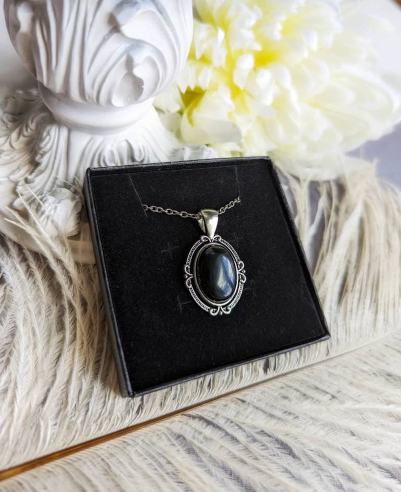 Black Obsidian Crystal Necklace - Silver Flourish Pendant Setting on a black display card, placed on a textured surface with flowers in the background.