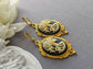 Pair of gothic black skull cameo drop earrings with detailed ivory lolita skull reliefs on a gray surface with white flowers.