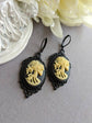 Pair of gothic black skull cameo drop earrings with detailed ivory lolita skull reliefs on a gray surface with white flowers in the background