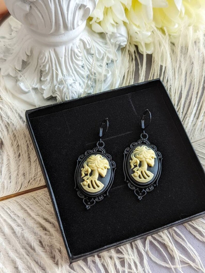 Pair of gothic black skull cameo drop earrings with detailed ivory lolita skull reliefs on a black display box with a decorative white background.