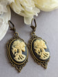 Pair of gothic black skull cameo drop earrings with detailed ivory lolita skull reliefs on a light gray surface with white flowers in the background.