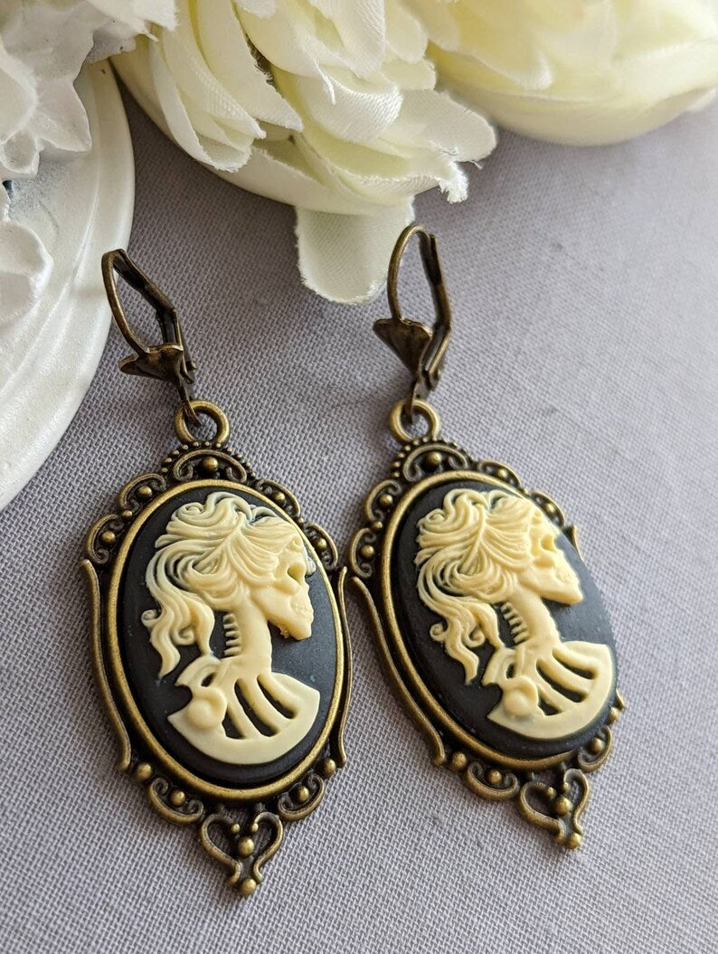 Pair of gothic black skull cameo drop earrings with detailed ivory lolita skull reliefs on a light gray surface with white flowers in the background.