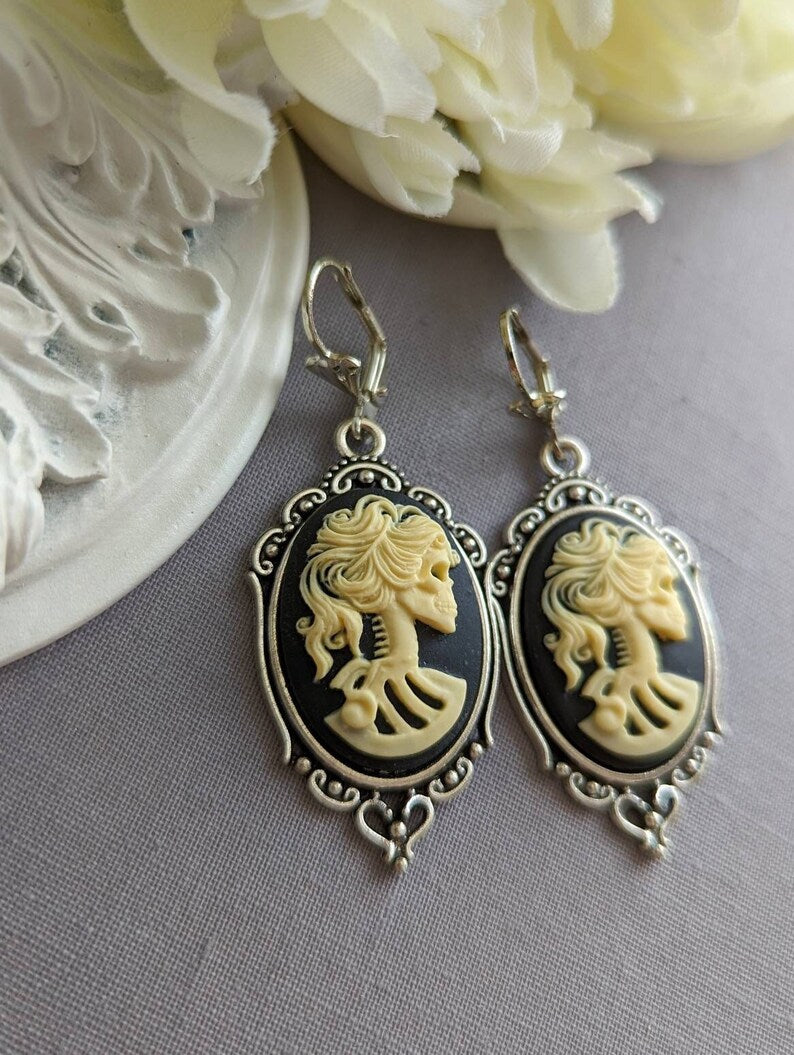Pair of gothic black skull cameo drop earrings with detailed ivory lolita skull reliefs on a gray surface with white flowers in the background.