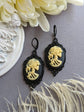 Pair of gothic black skull cameo drop earrings with detailed ivory lolita skull reliefs on a gray surface with flowers and feathers.