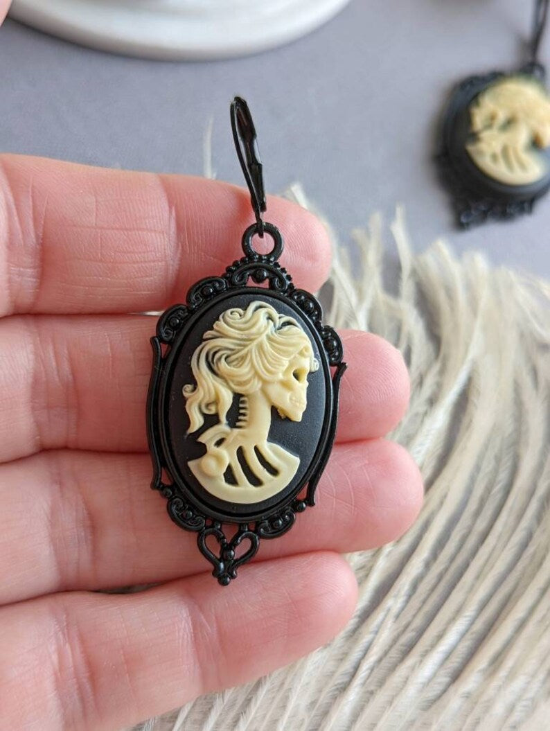 Pair of gothic black skull cameo drop earrings with detailed ivory lolita skull reliefs held in a hand with a blurred background
