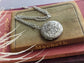 Silver round floral etched locket necklace by Papillion Era on a book with decorative elements