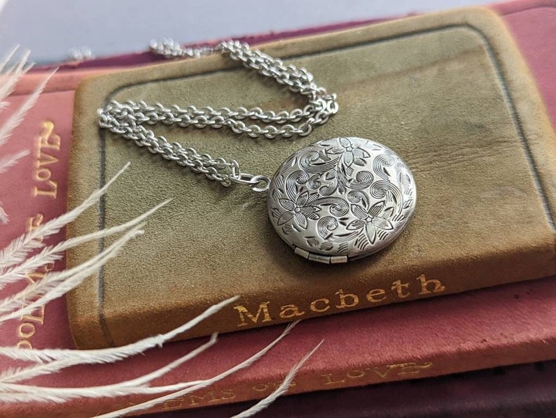 Silver round floral etched locket necklace by Papillion Era on a book with decorative elements