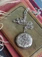 Silver round floral etched locket necklace by Papillion Era on an old book