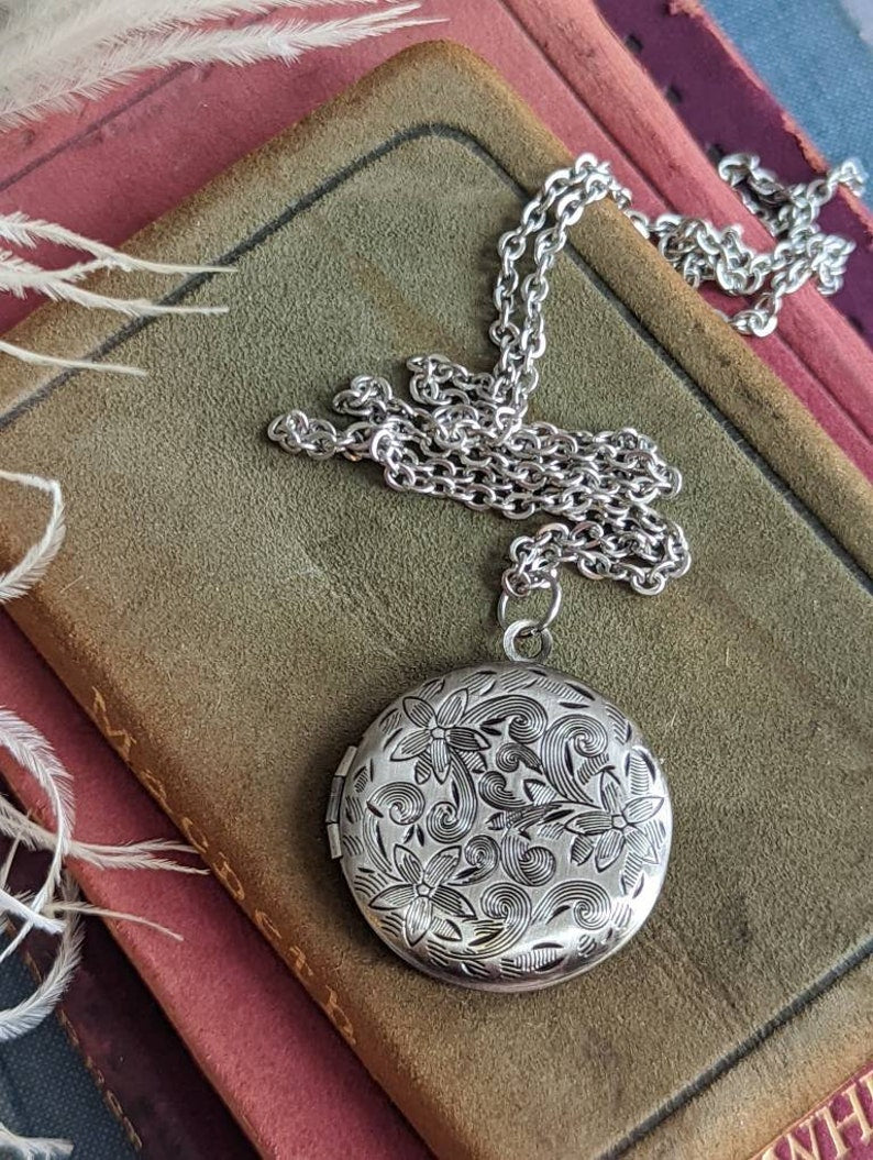 Silver round floral etched locket necklace by Papillion Era on an old book