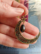 Antiqued Brass Filigree Crescent Moon Earring with Black Teardrop held between fingers with a blurred background