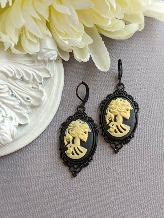 Pair of gothic black skull cameo drop earrings with detailed ivory lolita skull reliefs on a gray surface with white flowers in the background