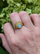 Art Deco Fan Ring with Iridescent Blue Opal - Gold Finish on a person's finger against a green leafy background