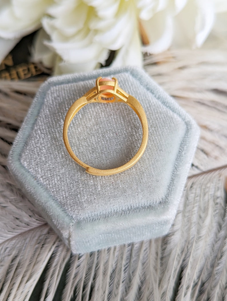 Side view of matte gold ring in a hexagonal light blue velvet box on a fluffy white surface with flowers in the background.