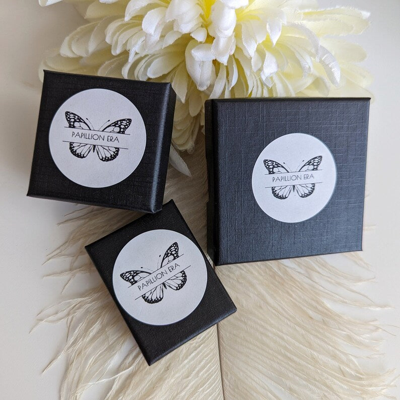 Three black boxes with white labels featuring a butterfly logo on a light wooden surface with white flowers in the background.