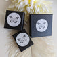 Three black boxes with white labels featuring a butterfly logo on a light wooden surface with white flowers in the background.