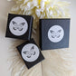 Three black boxes with white labels featuring a butterfly logo on a light wooden surface with white flowers in the background.