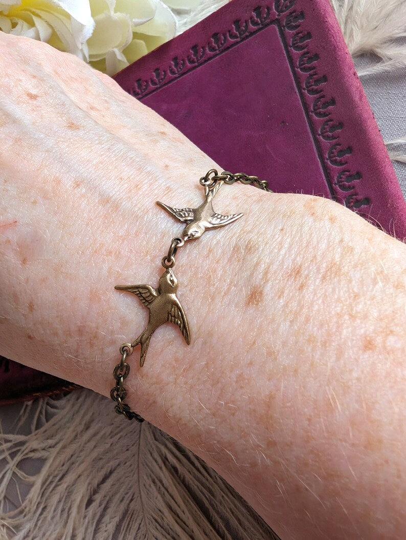 Dainty Swallow Bracelet: Antiqued Brass Bird Charm, Minimalist Jewelry on a wrist, with a purple book and flowers in the background