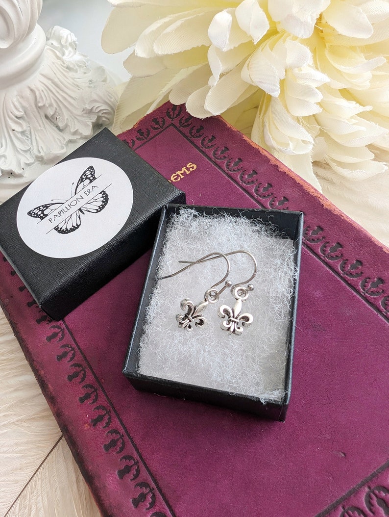 Fleur de Lis Earrings - Pewter French Lily Flower - Victorian Jewelry in a jewelry box with a brand logo on a decorative surface.