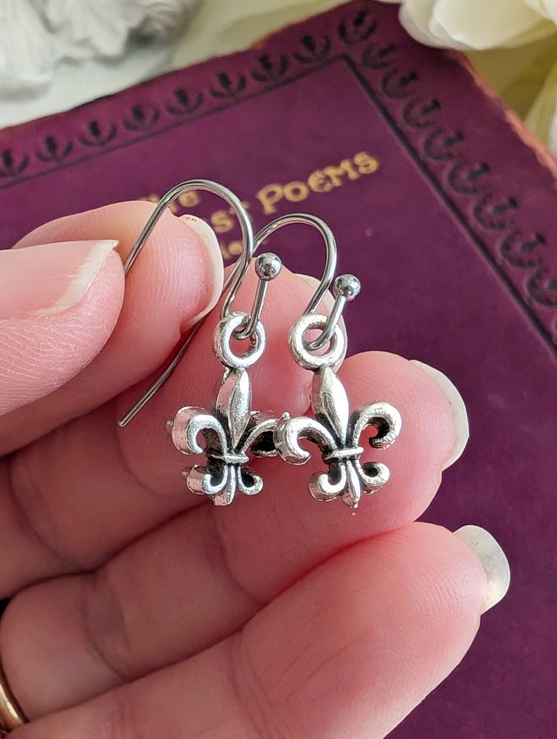 Silver fleur-de-lis earrings held between fingers with a purple book in the background