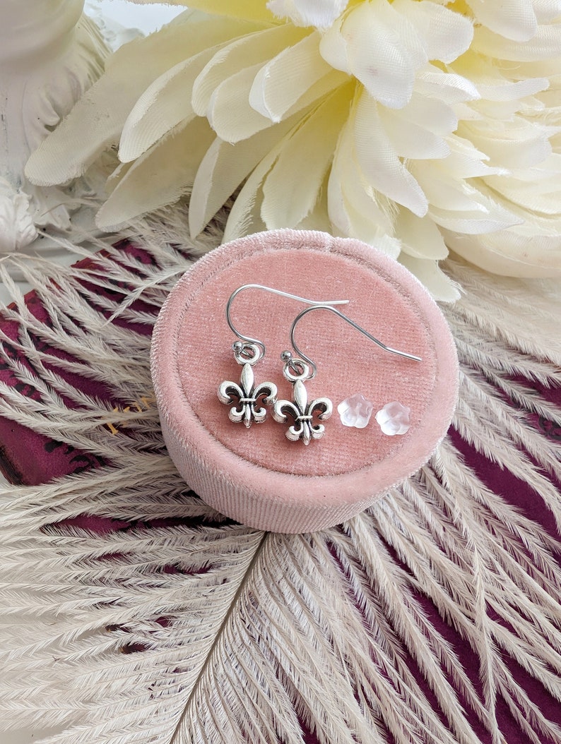Silver fleur de lis earrings and silicone earring backings displayed on a pink velvet box with white flowers and feathers