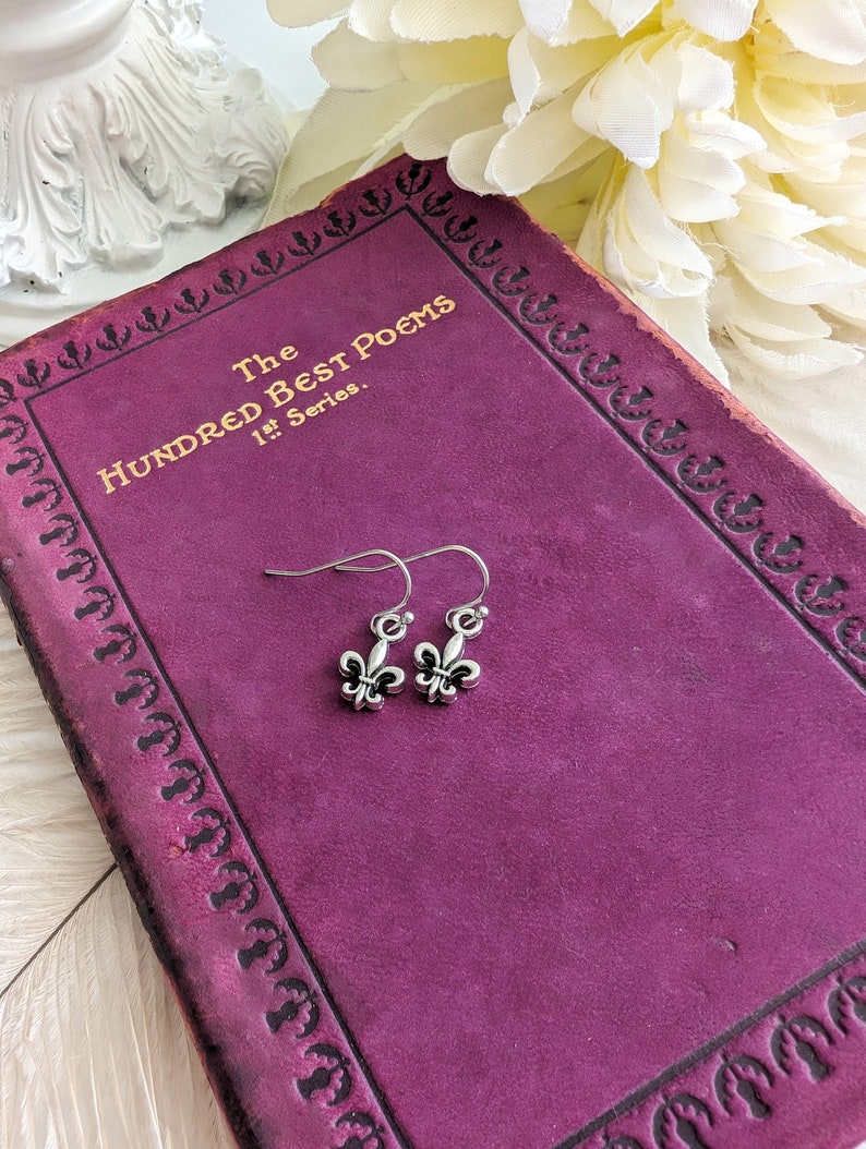 Fleur de Lis Earrings - Pewter French Lily Flower - Victorian Jewelry displayed on a purple poetry book, surrounded by white flowers.