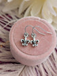 Silver fleur-de-lis earrings on a pink velvet cushion with white flowers in the background