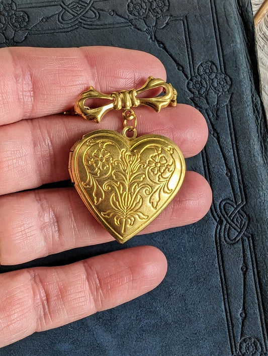 Gold-plated heart-shaped locket brooch with beautiful etched design on the locket face. Bow design pin. Shown held between fingers against a textured dark background