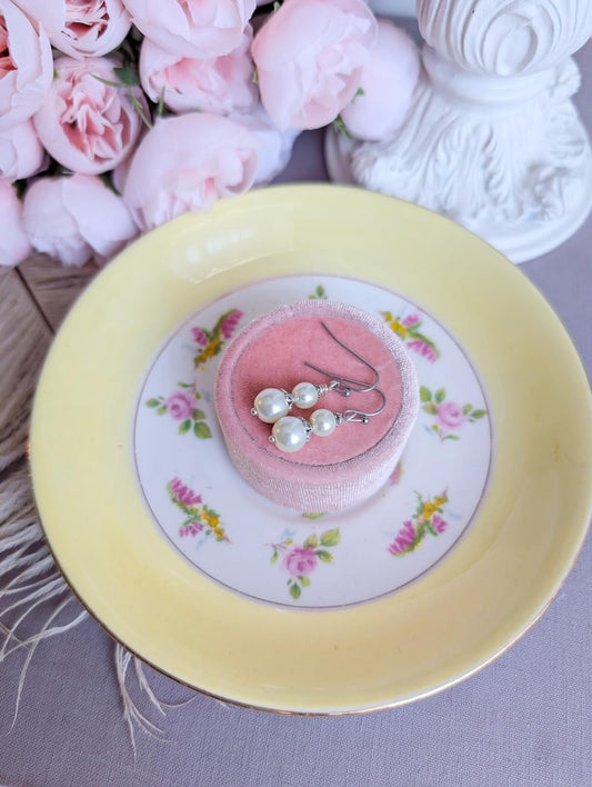 Pair of ivory double pearl drop earrings with antiqued silver filigree bead caps, on a decorative plate with flowers in the background
