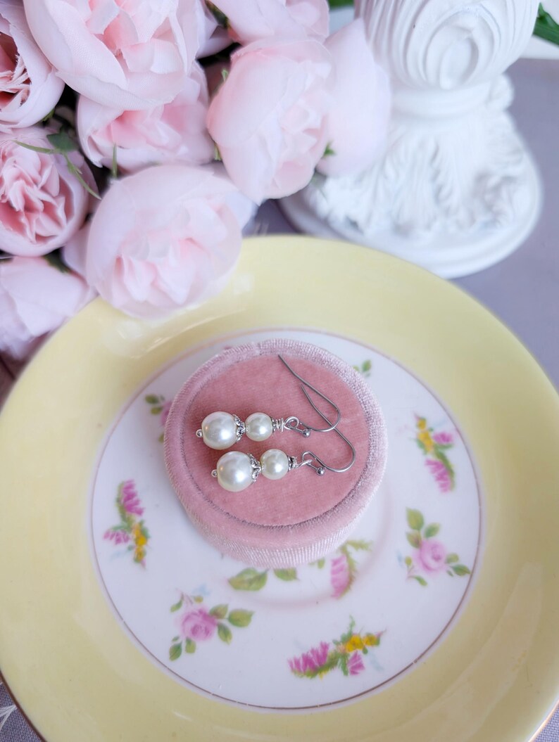 Pair of ivory double pearl drop earrings with antiqued silver filigree bead caps on a pink jewelry box with pink flowers in the background