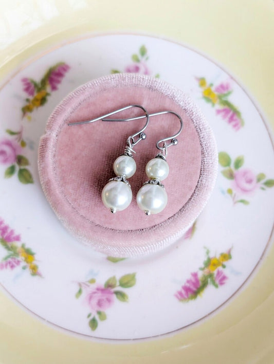 Pair of ivory double pearl drop earrings with antiqued silver filigree bead caps. on a pink jewelry box with a floral-patterned plate underneath